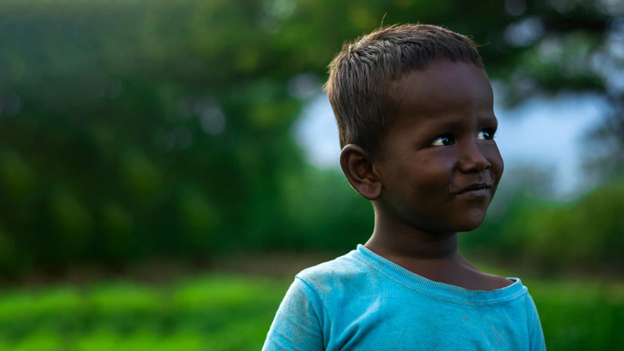 A young boy in a faded blue T-shirt smiles and glances to one side, against a blurred green field and trees.