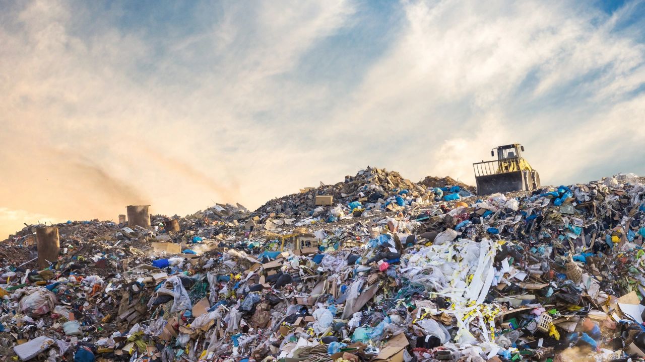 A yellow bulldozer on top of a vast landfill heap of plastic bags, paper and rubbish, under a cloudy sky lit by a low sun.