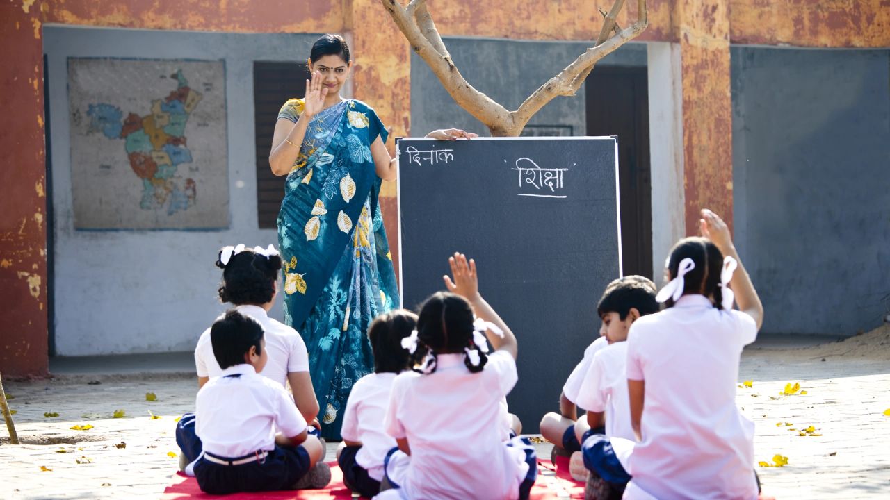 A teacher in a blue floral sari stands by a blackboard headed ‘शिक्षा’ (education) in a schoolyard, raising her hand, while children in white uniforms sit on a red mat in front of her, some with their hands raised.