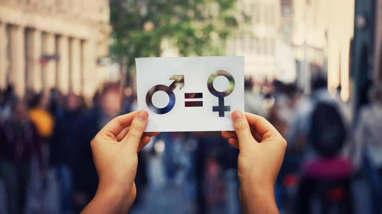 Hands holding up a paper card on a busy street, with the male and female symbols joined by an equals sign.