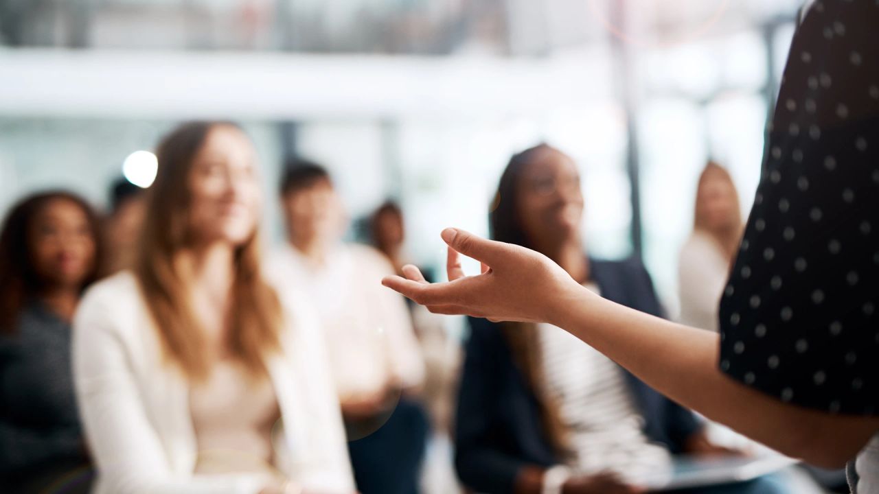 A speaker in a polka-dot top gestures with an open hand towards a blurred audience of young women seated in rows, who listen and smile.