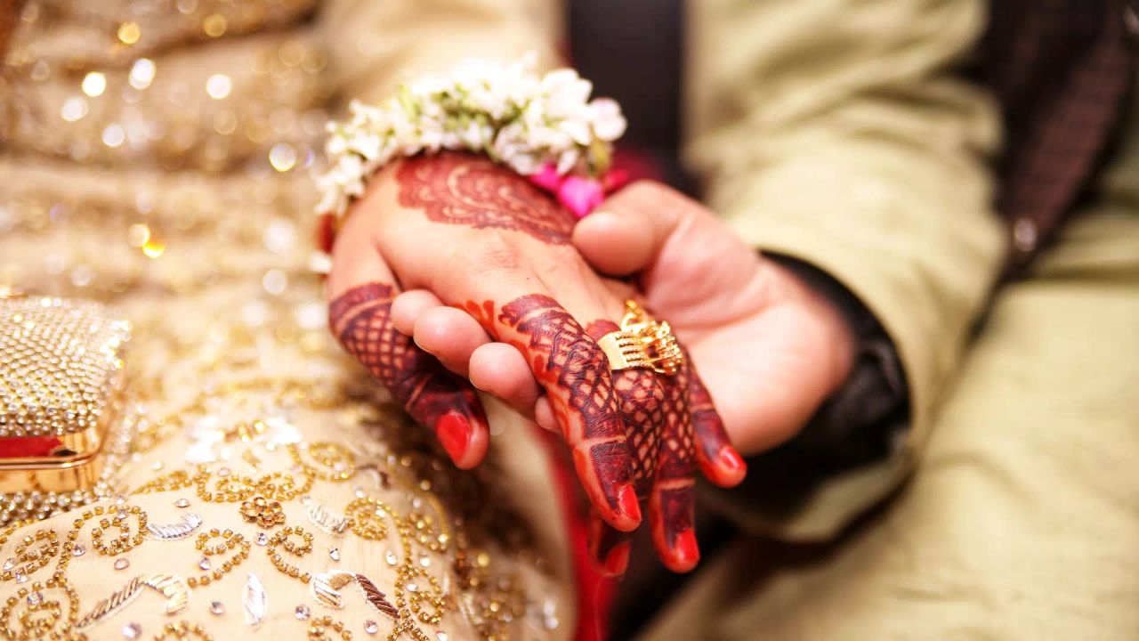 A bride’s hennaed hand, with a flower bracelet and a gold ring, resting in a groom’s hand.