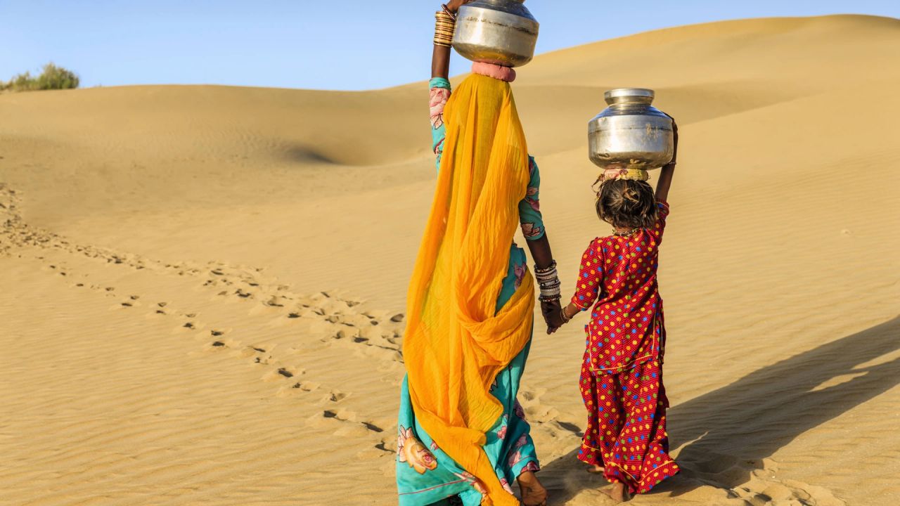 A woman in a yellow and turquoise sari and a young girl in a red polka-dot dress walk hand in hand across desert sand dunes, each balancing a metal water pot on her head.